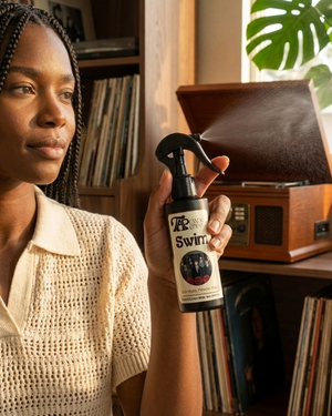 Woman holding a spray bottle labeled Swim in a room with a record player and books.